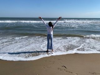 Person standing on a beach with arms raised, facing the ocean waves under a clear blue sky.